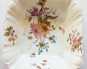May include: A white porcelain square dish with a scalloped edge. The dish is decorated with a floral pattern in shades of pink, yellow, green, and blue.