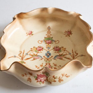May include: A cream-colored ceramic bowl with a scalloped edge and gold trim. The bowl is decorated with a floral pattern in shades of pink, blue, and green, and features two birds in flight.