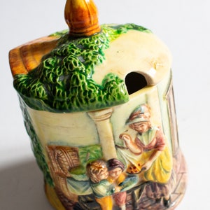 May include: A ceramic sugar bowl with a green and yellow lid shaped like a house. The bowl is decorated with a scene of people in a village setting.