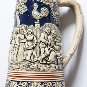 May include: A ceramic beer stein with a blue and white design. The stein features a rooster, a fence, and people in traditional clothing. The handle is shaped like a tree branch.