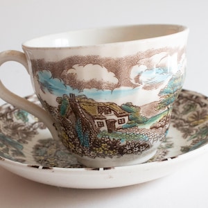 May include: A vintage teacup and saucer set. The cup and saucer feature a landscape scene with a cottage, trees, and clouds in shades of brown, blue, and green. The cup has a curved handle and sits on a matching saucer.