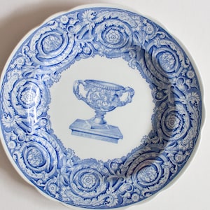 May include: A white ceramic plate with a blue floral and scrollwork border. The centre features a blue illustration of a decorative urn on a pedestal. The plate has a scalloped edge.