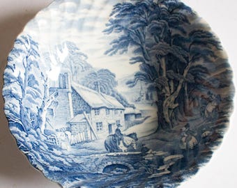 May include: A blue and white ceramic plate with a scalloped edge. The plate features a detailed landscape scene with a cottage, trees, and figures on horseback. The design is reminiscent of classic transferware.