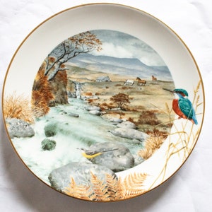 May include: A white ceramic plate with a gold rim features a watercolor painting of a river flowing through a mountainous landscape. The scene includes a small waterfall, rocks, trees, and two birds, one perched on a branch and the other on a rock.