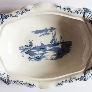 Puede incluir: Un plato ovalado blanco para servir con un diseño floral azul y blanco alrededor del borde. El centro del plato presenta una escena azul y blanca de un molino de viento y veleros en una masa de agua.