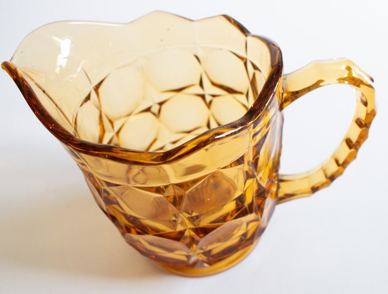 May include: Amber glass pitcher with a faceted design and a handle. The pitcher has a wide mouth and a rounded base.