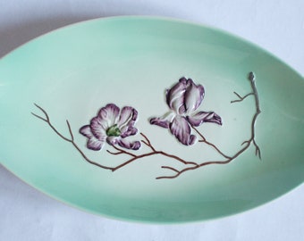 May include: A light green oval serving dish with a raised design of two purple flowers and a silver branch.