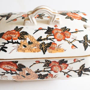 May include: A white ceramic serving dish with a lid, decorated with orange, black, and gold floral patterns. The rectangular dish has a handle on the lid and side handles. The design features blossoms and branches.
