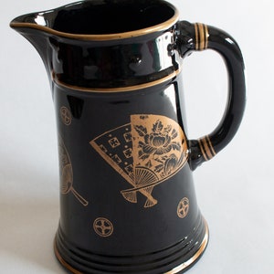 May include: Black ceramic pitcher with a gold rim and handle accents. The pitcher features gold fan and floral designs. The pitcher has a spout for pouring and a curved handle for easy grip. A decorative piece for serving beverages.