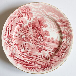 May include: A pink and white ceramic plate featuring a detailed illustration of a rural scene. The scene depicts a cottage, trees, and farm animals. The plate has a gold rim.