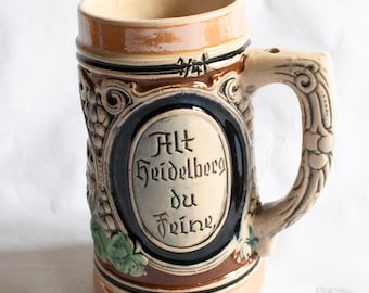 May include: A brown and white ceramic beer stein with a blue and white oval design that says "Alt Heidelberg du Feine". The stein has a handle with a sculpted design.