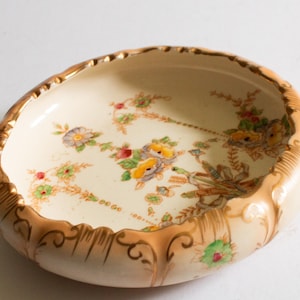 May include: A cream-coloured ceramic bowl with a gold rim and floral pattern. The bowl has a scalloped edge and is decorated with delicate flowers and leaves in shades of yellow, red, and green.