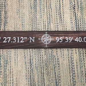 May include: A wooden sign with a compass design and the coordinates "36° 8' 27.312" N  95° 39' 40.032" W" in white lettering.