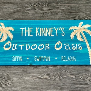 May include: A rectangular wooden sign painted turquoise with white text and palm tree designs. The text reads "THE KINNEY'S OUTDOOR OASIS" with the words "SIPPIN • SWIMMIN • RELAXIN" below.