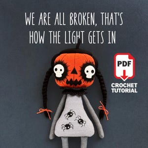 May include: A handmade crochet doll with an orange pumpkin head featuring white skull eyes and a black stem. The doll wears a gray dress with black spider accents and long black braids. Text reads "WE ARE ALL BROKEN, THAT'S HOW THE LIGHT GETS IN" and "CROCHET TUTORIAL".