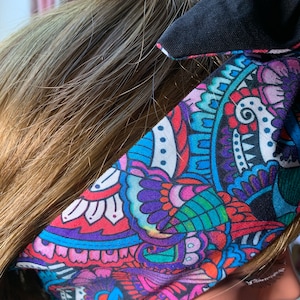 Op de afbeelding: Een kleurrijk hoofdband met een paisley-patroon in tinten blauw, roze, paars en groen. Het hoofdband is gemaakt van stof en heeft een zwarte rand.