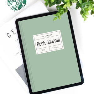 May include: A green digital tablet with a black frame displaying a book journal cover. The cover has a white label with the text "Notebook Book Journal" and the dates "2021" and "February".