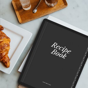 May include: A black digital recipe book with the text "Recipe Book" in white on the cover. The book is on a wooden surface with a glass of water and a cup of coffee.