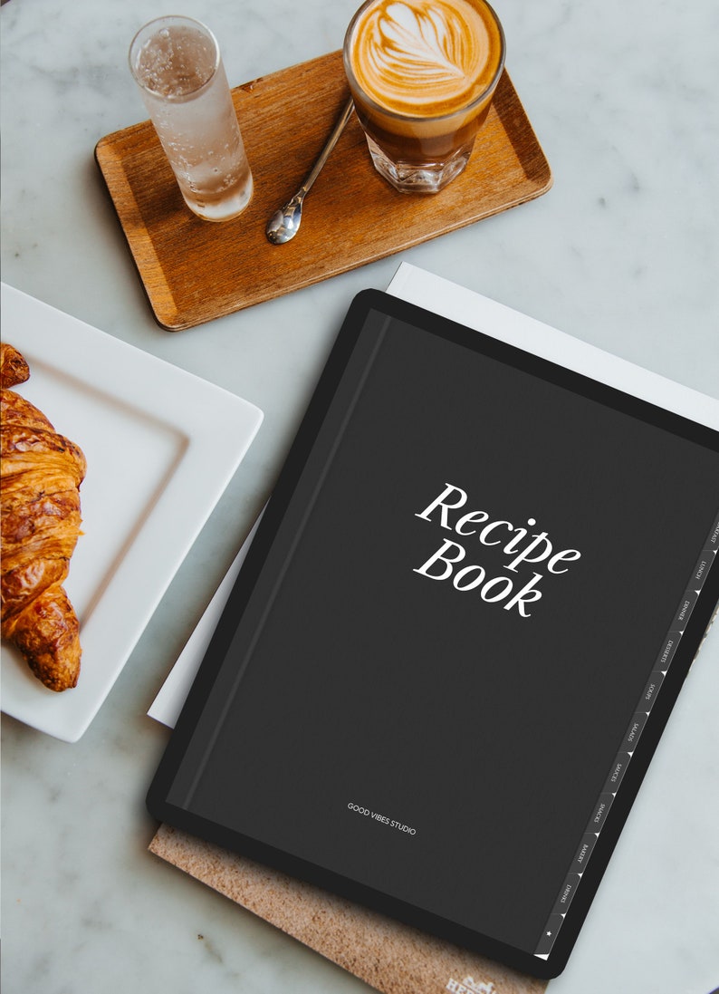 May include: A black digital recipe book with the text "Recipe Book" in white on the cover. The book is on a wooden surface with a glass of water and a cup of coffee.