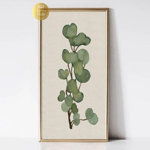May include: A framed botanical print showing a watercolour eucalyptus branch with green leaves and brown stems. The artwork is set against a beige background and framed in a gold-coloured frame. A gold sticker is in the upper left corner.