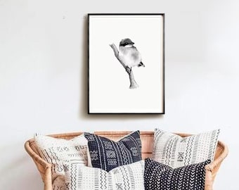 Op de afbeelding: Een zwart-witte tekening van een vogel die op een tak zit, ingelijst in een zwart frame en hangend aan een witte muur boven een rieten bank met zwart-witte gepatroneerde kussens.