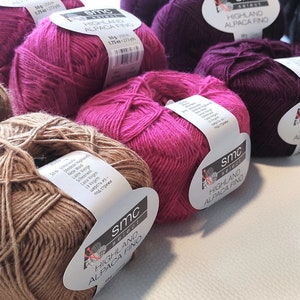 Op de afbeelding: Een verzameling garenbollen in verschillende kleuren, waaronder bruin, roze en paars. Het garen is geëtiketteerd met "Highland Alpaca Fino" en "SMC Yarns".