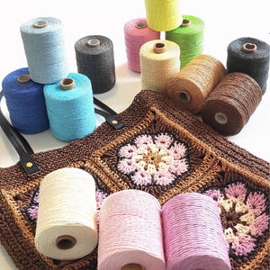 Può includere: Una varietà di rocchetti di filo di corda di cotone colorati, alcuni sono impilati uno sopra l'altro, altri sono appoggiati su una borsa di tela a crochet marrone. La borsa è realizzata con un motivo di grandi fiori.