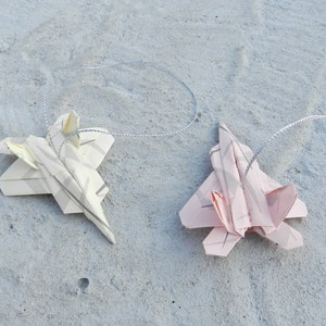 Könnte beinhalten: Zwei Origami-Papierflugzeuge hängen an einer silbernen Schnur. Ein Flugzeug ist weiß mit grauen Streifen und das andere ist rosa mit grauen Streifen.