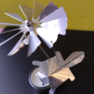 May include: A silver candle carousel with a floral design. The carousel has a spinning top with petal-shaped blades and a lower element with a similar design. The candle sits in a small, round holder.