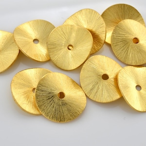 May include: A selection of gold-coloured, irregularly shaped metal discs with a radial brushed texture. Each disc features a small, central hole. The discs are scattered on a white surface, creating a bright and reflective display.