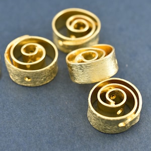 Op de afbeelding: Vijf gouden metalen ringen met een spiraalvormig ontwerp. De ringen hebben een getextureerd oppervlak en zijn gerangschikt op een blauwe achtergrond.