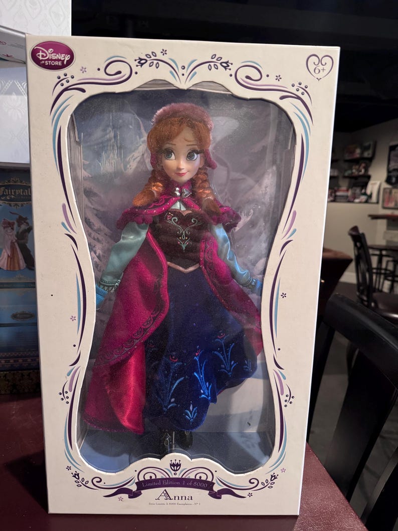 May include: A Disney Store Anna doll in a decorative box. The doll has red hair and wears a blue and purple dress with a pink cape. The box is white with a decorative border and the text "Anna".