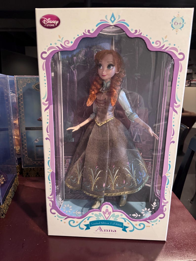 May include: A Disney Store Anna doll in a display box. The doll has red hair and wears a brown and blue dress. The box is white with a purple and teal design and the text "Anna" and "Limited Edition 1 of 5000".