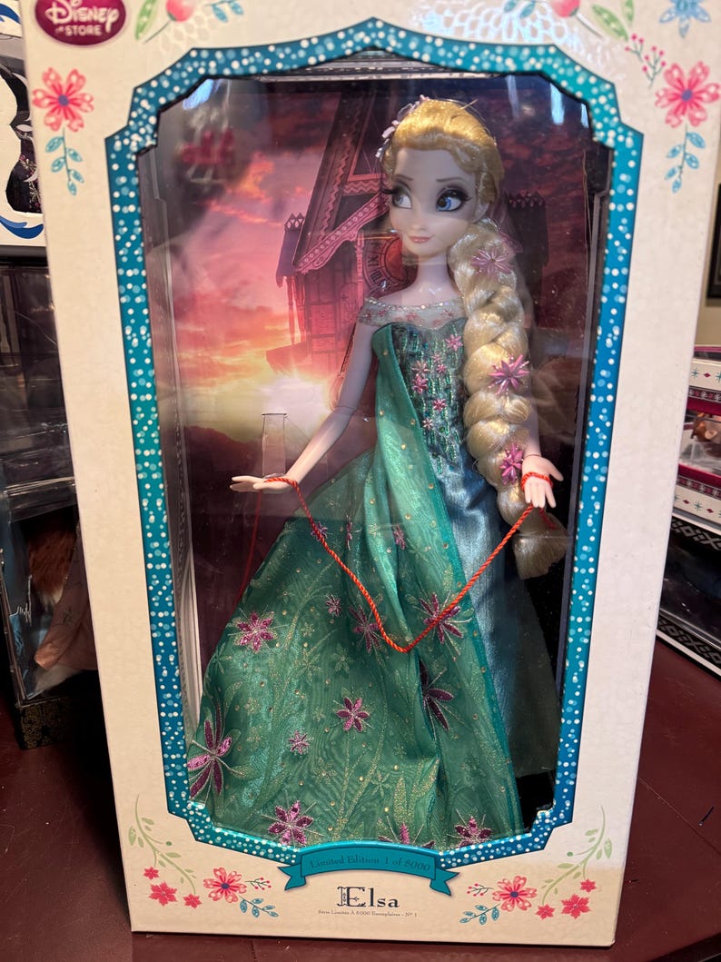 May include: A Disney Store Elsa doll in a display box. The doll has blonde hair in a long braid adorned with pink flowers. She wears a teal and green gown with floral accents. The box features a scenic background and the name "Elsa".