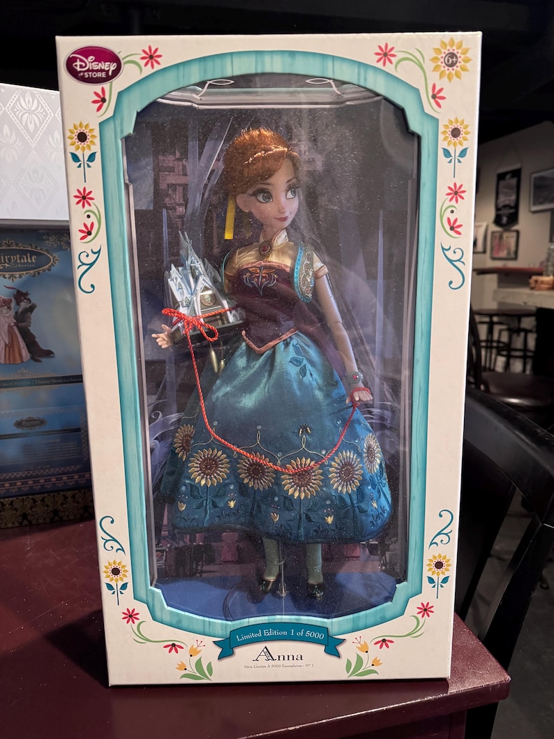 Op de afbeelding: Een Disney Store Anna-pop in een decoratieve doos. De pop draagt een turquoise jurk met zonnebloemaccenten en een bordeauxrood lijfje. De doos is wit met bloemen en turquoise accenten, met de tekst "Limited Edition 1 of 5000".