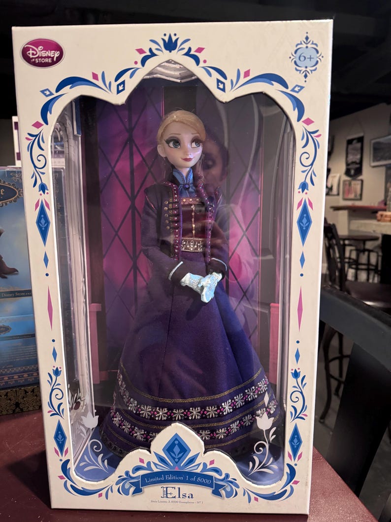 K&ouml;nnte beinhalten: Eine Elsa-Puppe aus dem Disney Store in einer Box. Die Puppe tr&auml;gt ein lila-schwarzes Kleid mit goldenen Akzenten. Die Box ist wei&szlig; mit blauen und lila dekorativen Elementen und dem Disney Store-Logo. Der Text "Limited Edition 1 of 5000" ist sichtbar.
