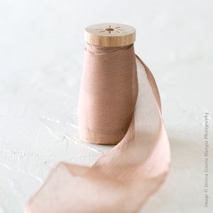 May include: A spool of light brown silk ribbon with a wooden spool. The ribbon is unfurled and draped over a white surface.