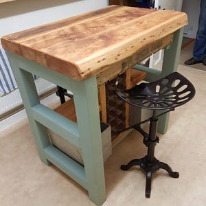 May include: A high-top table with a thick, natural-edge wood grain top and sage green painted legs. The base features a built-in wine rack and a metal storage bin. Beside the table stands a black metal tractor-style stool with a swivel seat.