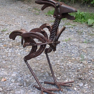 K&ouml;nnte beinhalten: Rostige Metallskulptur eines Hahns, gefertigt aus Hufeisen und Metallteilen. Der Hahn hat einen Federhals und steht auf einem Kiesboden. Ein einzigartiges, rustikales Kunstwerk, das handwerkliches Geschick zeigt.