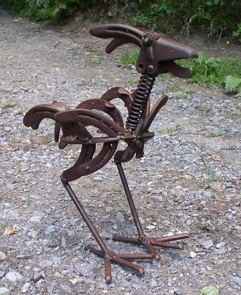K&ouml;nnte beinhalten: Rostige Metallskulptur eines Hahns, gefertigt aus Hufeisen und Metallteilen. Der Hahn hat einen Federhals und steht auf einem Kiesboden. Ein einzigartiges, rustikales Kunstwerk, das handwerkliches Geschick zeigt.