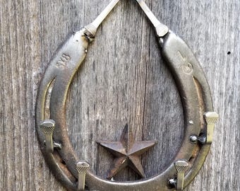 May include: Rustic metal horseshoe wall decor with a star accent. The horseshoe is mounted with nails and has two metal pieces attached at the top, joined by a screw. The background is weathered wood.