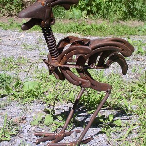 K&ouml;nnte beinhalten: Eine rostige Metallskulptur eines Hahns, hergestellt aus Hufeisen und Federn. Die Skulptur hat einen detaillierten Kopf, K&ouml;rper und Beine mit einem Federnhals. Der Hahn steht auf einem Bett aus Kies und Gras und hat ein nat&uuml;rliches, verwittertes Aussehen.