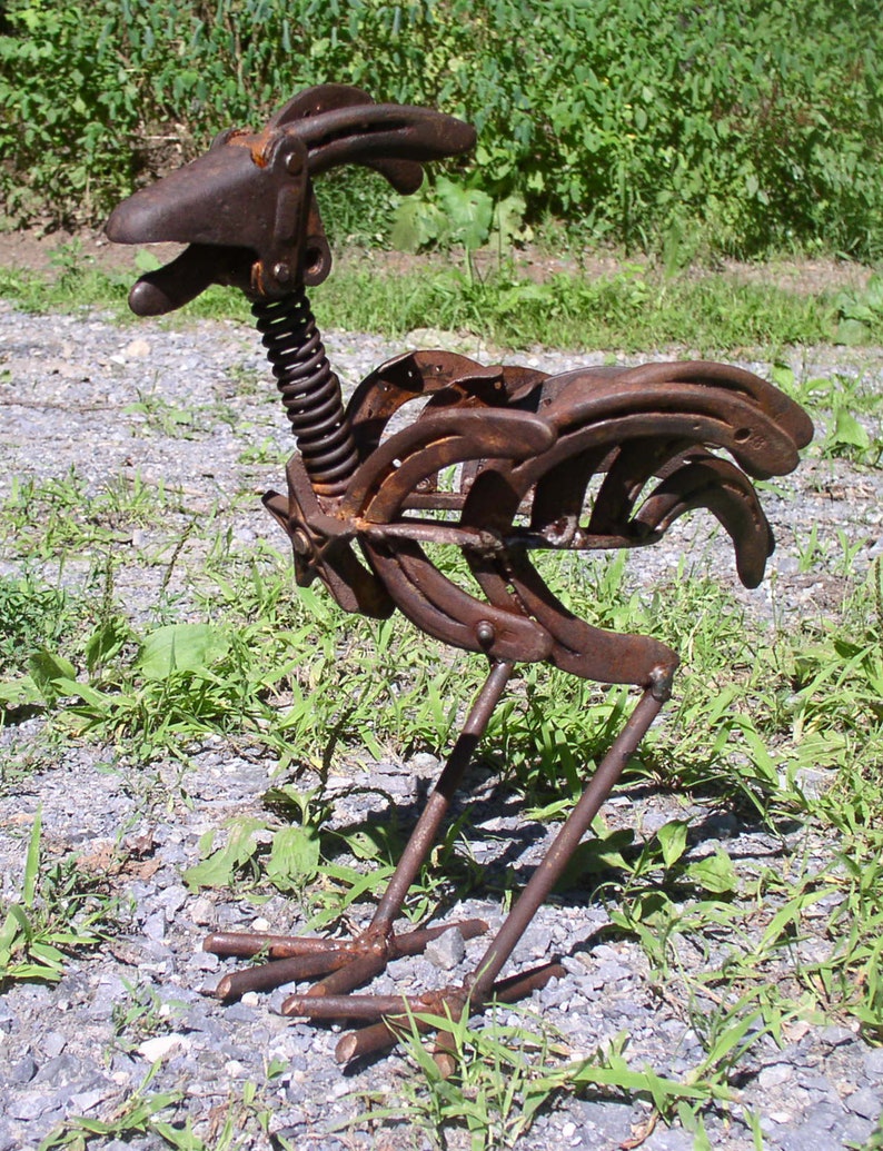 K&ouml;nnte beinhalten: Eine rostige Metallskulptur eines Hahns, hergestellt aus Hufeisen und Federn. Die Skulptur hat einen detaillierten Kopf, K&ouml;rper und Beine mit einem Federnhals. Der Hahn steht auf einem Bett aus Kies und Gras und hat ein nat&uuml;rliches, verwittertes Aussehen.