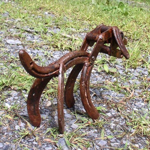 May include: A rusty metal sculpture of a dog made from horseshoes. The sculpture is standing on a bed of gravel and grass.