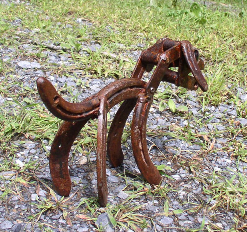 May include: A rusty metal sculpture of a dog made from horseshoes. The sculpture is standing on a bed of gravel and grass.
