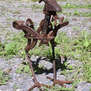 K&ouml;nnte beinhalten: Eine rostige Metallskulptur eines Vogels, hergestellt aus recycelten Metallteilen. Der Vogel hat einen Federmechanismus f&uuml;r den Hals, ein sternf&ouml;rmiges Element und Hufeisenfl&uuml;gel. Die Skulptur steht auf einem Kiesboden mit gr&uuml;nem Gras.