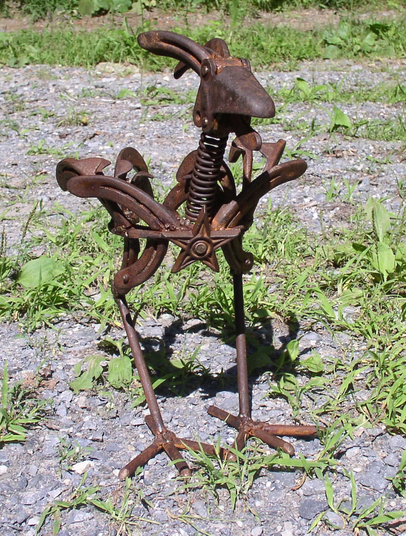 K&ouml;nnte beinhalten: Eine rostige Metallskulptur eines Vogels, hergestellt aus recycelten Metallteilen. Der Vogel hat einen Federmechanismus f&uuml;r den Hals, ein sternf&ouml;rmiges Element und Hufeisenfl&uuml;gel. Die Skulptur steht auf einem Kiesboden mit gr&uuml;nem Gras.