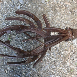 K&ouml;nnte beinhalten: Ein rostiges Metall-Gartenwerkzeug mit gebogenen Zinken und einem Federmechanismus. Das Werkzeug dient zum Auflockern des Bodens und zur Vorbereitung der Bepflanzung. Das Bild ist von oben aufgenommen und zeigt das Werkzeug vor einem Kieshintergrund.