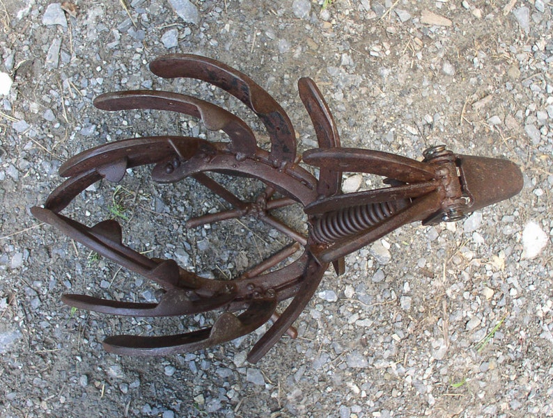 K&ouml;nnte beinhalten: Ein rostiges Metall-Gartenwerkzeug mit gebogenen Zinken und einem Federmechanismus. Das Werkzeug dient zum Auflockern des Bodens und zur Vorbereitung der Bepflanzung. Das Bild ist von oben aufgenommen und zeigt das Werkzeug vor einem Kieshintergrund.