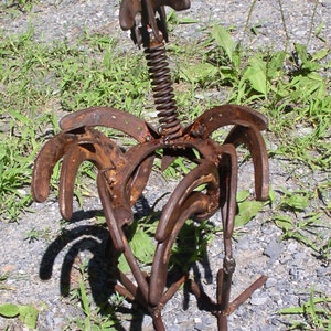 K&ouml;nnte beinhalten: Eine rostige Metallskulptur eines Vogels, gefertigt aus Hufeisen und einer Feder. Die Skulptur hat eine verwitterte, braune Oberfl&auml;che und steht auf Kies und Gras. Der Vogelk&ouml;rper wird durch gebogene Hufeisen geformt.